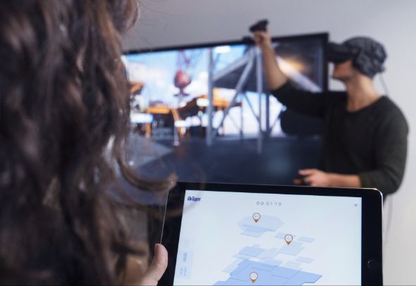 An over the shoulder photo of a person holding an ipad for controlling the VR game, in the background a person with VR glasses playing